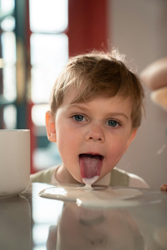 Adorable young boy playfully licking a milk spill from a table indoors.